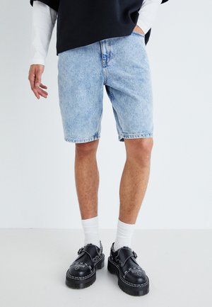Pantaloni scurți din denim albastru deschis, cu o croială lejeră, margine dantelată și design clasic cu cinci buzunare, asortați cu pantofi negri și șosete albe.