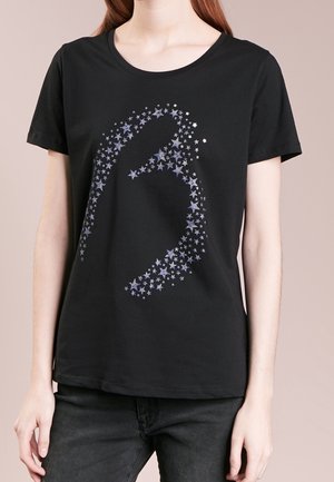 Czarny T-shirt z krótkim rękawem z dużym srebrnym wzorem gwiazdy tworzącym literę „B” na przodzie, noszony z czarnymi spodniami.