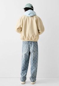 Beżowy oversize'owy sweatshirt z jasnoniebieskim kapturem, zestawiony z wzorzystymi jasnoniebieskimi dżinsami z geometrycznym wzorem. Dodatkowo akcentowany zieloną czapką.