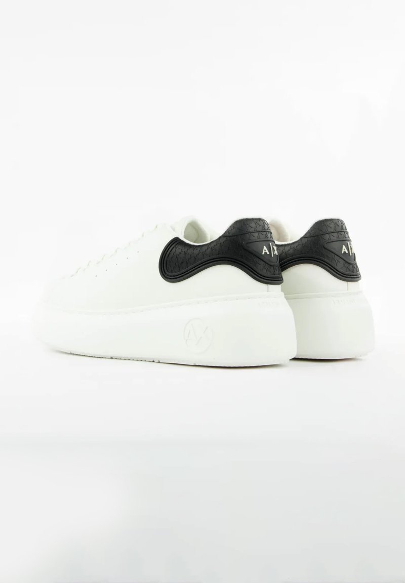 A fehér bőrből készült sportcipők sarkán fekete, texturált díszítés található, lekerekített orral és gumitalppal, amely sima felületű és logó díszítést tartalmaz.
