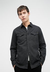 Giovane uomo con i capelli castani corti, che indossa una camicia nera con bottoni e tasche sul petto, in piedi con le mani nelle tasche davanti a uno sfondo semplice.