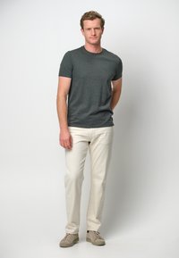 Mörkgrå t-shirt med kort ärm i kombination med krämfärgade raka byxor och ljusgrå sneakers, stående mot en neutral bakgrund.