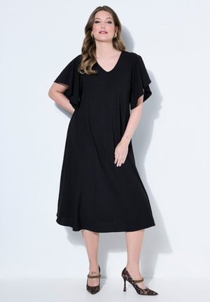 Femme debout portant une robe midi noire à col en V avec des manches volantées et des talons hauts imprimés léopard avec des brides de cheville en chaîne.