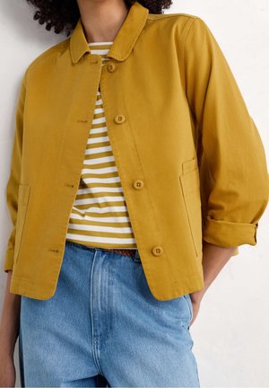 Femme portant une veste moutarde à boutons par-dessus une chemise rayée blanche et moutarde, avec un jean bleu clair, debout avec une main dans la poche.