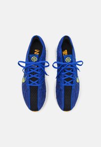 Blaue Sportschuhe mit einem strukturierten Strick-Obermaterial, schwarzen Akzenten, gelbem Logo und flachen Schnürsenkeln, die ein niedriges Profil und eine gepolsterte Sohle aufweisen.