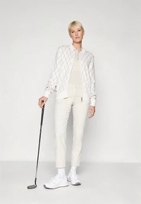 Witte bomberjas met een diamantpatroon, beige poloshirt, crèmekleurige broek en witte schoenen. Model houdt een golfclub vast.