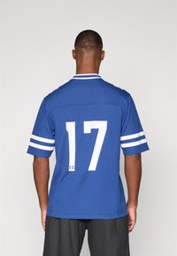 Blå kortermet jersey med hvite striper, med et stort hvitt tall "17" på baksiden og en kontrasterende kragedesign.