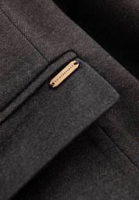 Tela gris oscuro con un tejido texturizado. Presenta una etiqueta de metal con el texto "QUINTESSENCE" asegurada con hilo morado en la solapa.