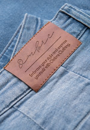 Sininen farkkukangas, jossa on näkyvä ruskea nahkainen etiketti, jossa on kohokuvioitu teksti. Etiketissä on merkin nimi "Ombre" ja iskulause.