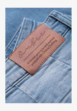 Blauwe spijkerstof met een opvallend bruin leren label met geperste tekst. Het label toont de merknaam "Ombre" en een slogan.