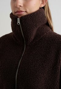 Bruine fleece zip-up jas met een hoge kraag, voorzien van een textuurstof en een zilveren rits, ideaal voor warmte en comfort.