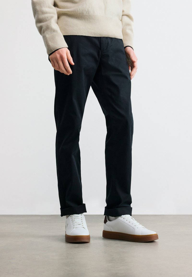 Άνδρας που φοράει μαύρο παντελόνι γυρισμένο στους αστραγάλους, μπεζ πουλόβερ και άσπρα αθλητικά παπούτσια με καφέ σόλες, στέκεται σε λείο δάπεδο.