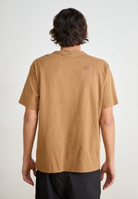 Braunes Baumwoll-T-Shirt mit kurzenÄrmeln, Rundhalsausschnitt und einem kleinen geprägten Logo auf dem oberen Rücken. Einfaches Design, lockere Passform.