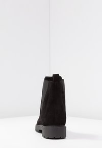 Botín de ante negro hasta el tobillo con tirador, paneles laterales elásticos y suela acanalada, mostrado desde la parte de atrás sobre una superficie y fondo blancos.