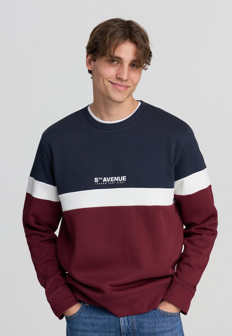 Giovane uomo con capelli castani che indossa una felpa blu navy, bianca e bordeaux con la scritta "5TH AVENUE UPPER EAST SIDE", mani nelle tasche dei jeans di denim.