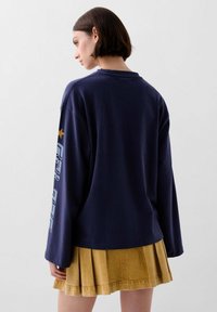 Camisa de manga larga azul marino con cuello redondo y texto impreso en la manga izquierda, combinada con una falda amarilla con pliegues. Tejido de algodón suave.