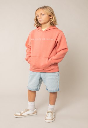 Jeune personne debout, mains dans les poches, portant un sweat à capuche corail marqué "DEELUXE ORIGINAL", un short en jean clair, des chaussettes blanches et des baskets.