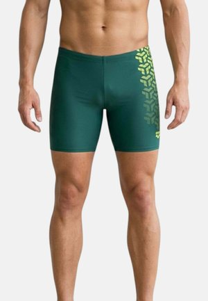 Uomo che indossa pantaloncini da bagno aderenti verde scuro con motivo geometrico giallo sulla coscia destra, in piedi con le braccia rilassate ai lati.