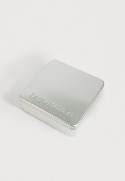 Mazs sudraba metāla trauks ar noapaļotām malām un uz vāka iegravētu "le gramme", novietots uz vienkārša baltā virsmas.