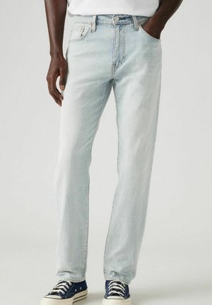 Homme portant un jean droit bleu clair, un t-shirt blanc et des baskets bleu marine avec des lacets blancs, debout les mains dans les poches.