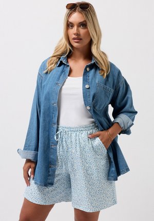 Mujer rubia con gafas de sol sobre la cabeza, camiseta sin mangas blanca, camisa vaquera oversize y pantalones cortos azules claros con flores y cordón, con la mano en el bolsillo.