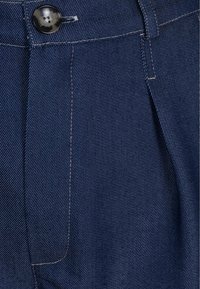 Marineblauwe denimbroeken met een knoopsluiting, opvallende stiksels en plooien voor extra detail en textuur.