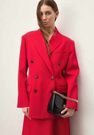 Raudonas dvieilis švarkas su prigludusia forma, juodais mygtukais ir šoninėmis kišenėmis. Derinamas su maža juoda rankine su auksiniu grandinėlės dirželiu.