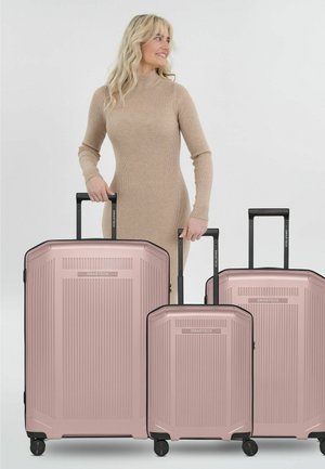 Blonde Frau in beigem Kleid hält die Griffe von drei rosa Hartschalenkoffern mit schwarzen ausziehbaren Griffen und Rollen.