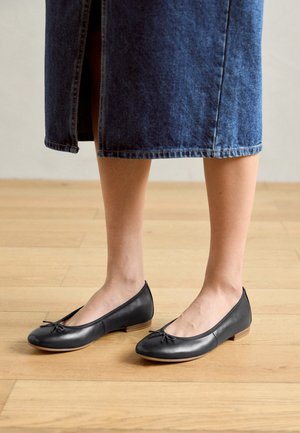 Czarne baleriny ze skóry z zaokrąglonym noskiem, małą kokardką i niskim drewnianym obcasem, zestawione z dżinsową spódnicą midi na drewnianej podłodze.