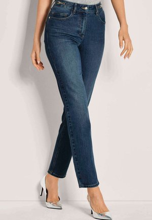 Frau in dunkelblauen schmalen Jeans und silbernen High Heels, die mit übereinander geschlagenen Beinen vor einem schlichten Hintergrund steht.