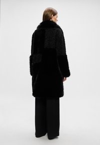 Melns mākslīgā ādas mētelis ar dažādām tekstūrām un rakstiem, ar augstu apkakli un platām piedurknēm. Uzvilkts pār melniem platām biksēm.