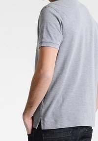 Szara bawełniana koszulka polo z krótkimi rękawami, płaskim kołnierzykiem, bocznymi rozcięciami i subtelną teksturą. Nosi ją osoba z rękami w kieszeniach.