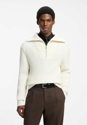 Ung mand iført en hvid ribstrikket sweater med halv lynlås over en sort skjorte og slips, kombineret med brune bukser og sort bælte, står mod en hvid baggrund.