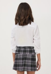 Camicia bianca a maniche lunghe con colletto, abbinata a una gonna a quadri nera con tocchi di viola e pieghe, vista da dietro.