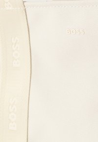 Tissu blanc avec un motif texturé arborant un logo "BOSS" en relief subtil. La sangle présente un tissage détaillé avec le nom de la marque.