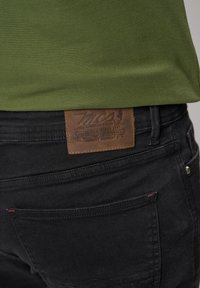 Jeans en denim noir avec une patch en cuir marron visible à la taille, comportant des détails de marque. Le tissu a une texture lisse.