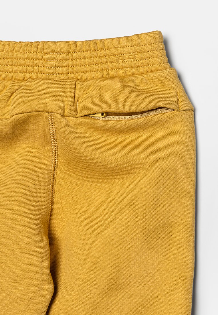 Pantalons de jogging jaunes avec une taille côtelée, une poche zippée sur le côté et une texture de tissu douce. Présente des coutures visibles et un design à coupe décontractée.