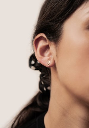 Nahaufnahme eines Frauenohrs mit einem kleinen goldfarbenen Ohrring in Form einer Meerjungfrau und dunklem Haar, das mit einem schwarz-weißen gepunkteten Haargummi zusammengebunden ist.