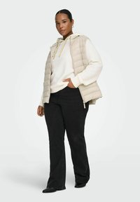 Beige puffervest over en kremfarget hettegenser, kombinert med svarte, vide jeans. Vesten har et horisontalt vattert design og glidelåslukking.