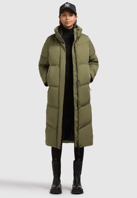 Pitkä oliivinvihreä untuvapuvun takki, jossa on korkea kaulus, napit ja sivutaskut. Mallilla on päällään mustat legginsit ja nilkkurit.