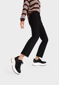 Sneakers nere chunky con tomaia testurizzata e suola bianca, abbinate a jeans neri cropped e una camicia a righe marroni a manica lunga.