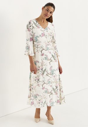 Vrouw die een witte midi-jurk met V-hals draagt, bedrukt met bloemen en vogels, met driekwartsmouwen met ruches, en beige puntige hakken, staand tegen een witte achtergrond.