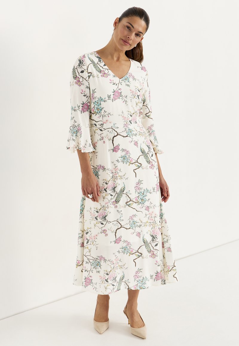 Kvinde iført en hvid V-hals midi-kjole med blomster- og fugleprint, trekvartlange ærmer med flæser og beige spidse hæle, stående mod en hvid baggrund.