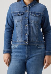 Jeansjacka i mellanblå denim med krage, två bröstfickor och knäppning med knappar. Långa ärmar med manschetter och en figursydd fåll.