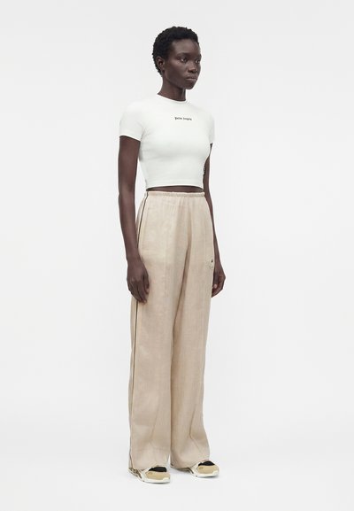 Μαύρη γυναίκα στέκεται, φοράει λευκό κοντό μπλουζάκι με μικρό κείμενο και φαρδιά μπεζ παντελόνια με μαύρες πλαϊνές ρίγες και μπεζ αθλητικά παπούτσια.