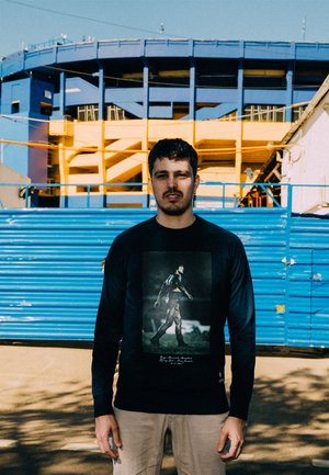 Zwarte lange mouwen shirt met een grafische print van een figuur die wandelt. De achtergrond bevat een levendige blauwe en gele stadionstructuur.
