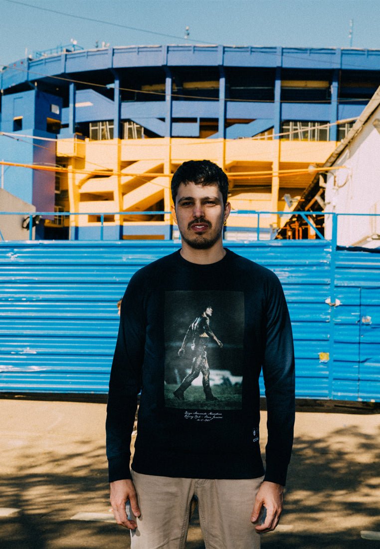 Zwarte lange mouwen shirt met een grafische print van een figuur die wandelt. De achtergrond bevat een levendige blauwe en gele stadionstructuur.