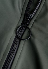 Primer plano de un tirador de cremallera negro y circular en una cremallera parcialmente cerrada con tela verde alrededor.