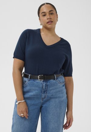 Žena nosi tmavomodrý top s výstrihom do V a krátkym rukávom, vložený do vysokých modrých džínsov s čiernym opaskom, pózujúca na jednolivom pozadí.