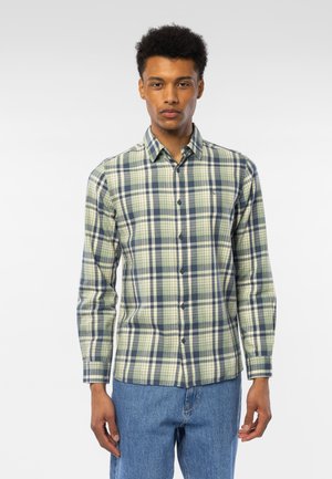 Ung mann som står, iført grønn og marineblå rutete knappeskjorte og blå jeans, mot en enkel lys bakgrunn.
